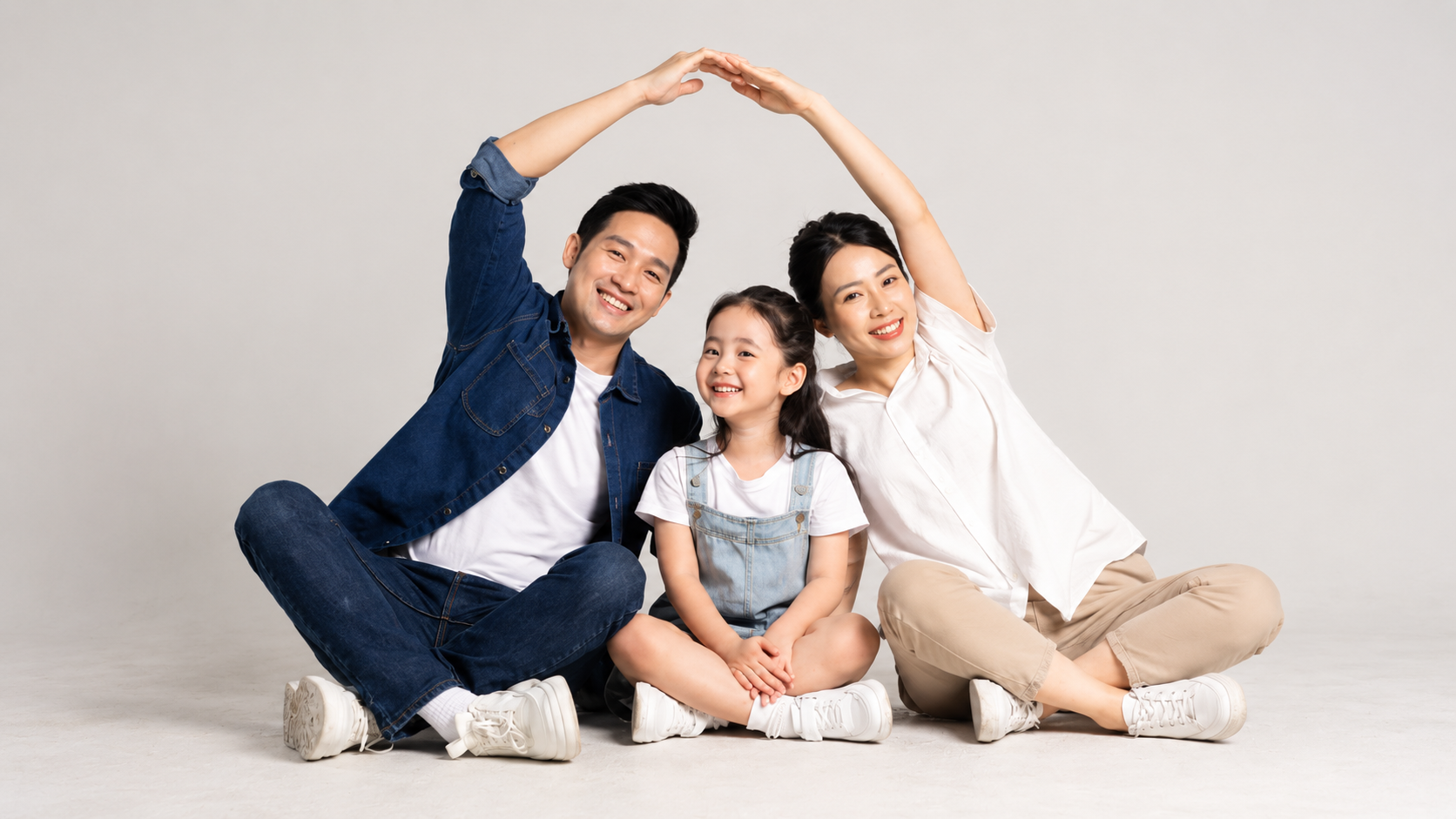 Smiling family of three sitting together on the floor, the parents making a roof shape with their arms over their daughter